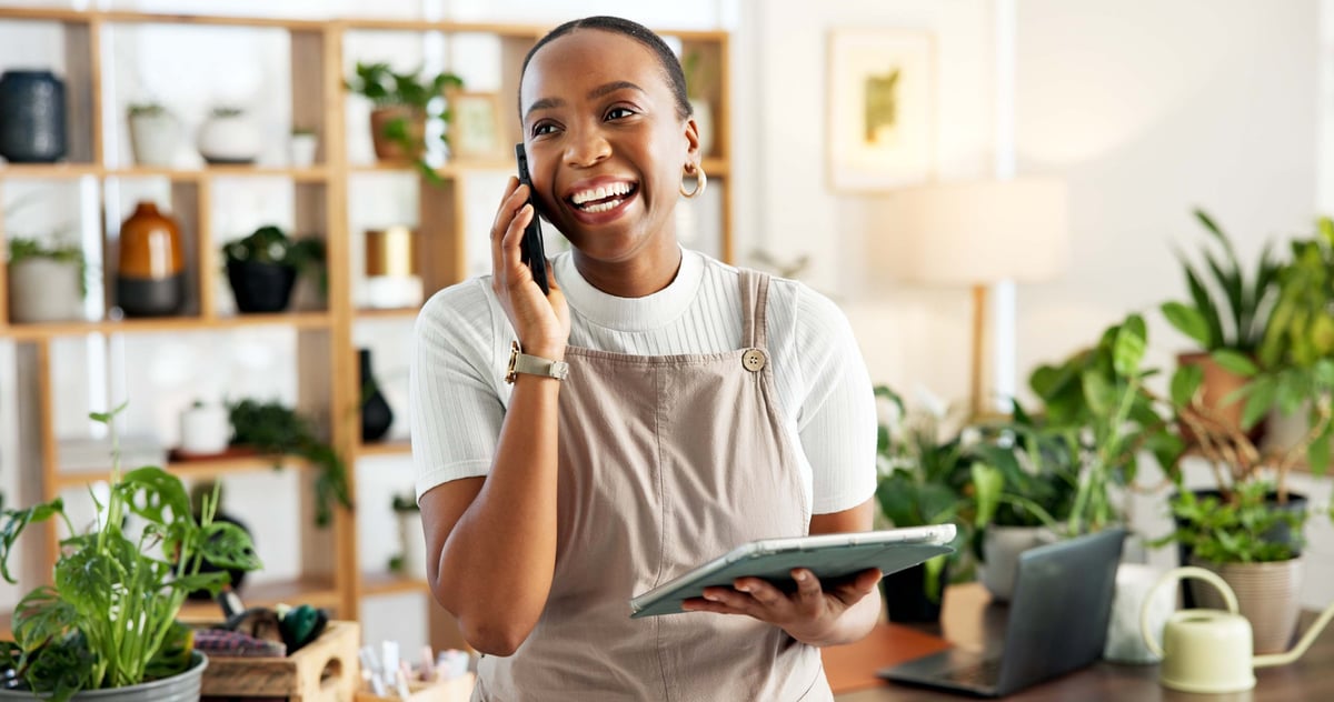 Black female florist smiling while using tablet and phone at her plant nursery shop